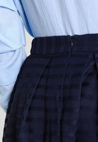 Detailní záběr světle modré košile zastrčené do plisované tmavě modré texturované sukně se zapínáním na zip na boku.