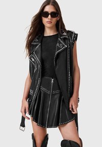 Zwarte leren mouwloze vest met studaccenten, gecombineerd met een bijpassende ritsrok. Model draagt zwarte zonnebril en heeft lang haar.