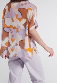 Chemise ample en tissu léger à motifs avec des formes géométriques orange, violettes et marron. Manches courtes, détail à plis dans le dos.