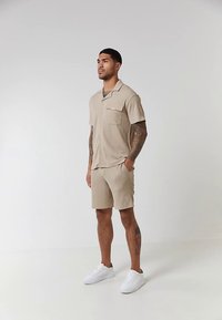Chemise beige à manches courtes avec une poche et un short assorti, en tissu texturé. Porté avec des baskets blanches, debout devant un fond uni.