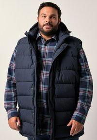 Námořnická izolační vesta s vysokým límcem a kapucí přes kostkovanou košili s dlouhým rukávem, která má modré, zelené a burgundské pruhy.