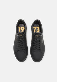Sapatilhas de couro preto com bico redondo, atacadores planos e logótipo da marca em relevo. Os detalhes em dourado exibem "1973" nas palmilhas.