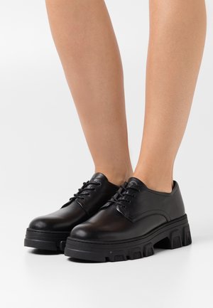 Chaussures en cuir noir avec une texture lisse, bout arrondi et épaisse semelle plateforme noire avec un motif géométrique. Design à lacets.