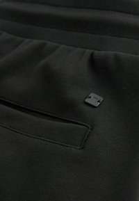 Gros plan sur un tissu noir avec un petit patch logo carré à côté d'un détail de poche horizontale cousue.