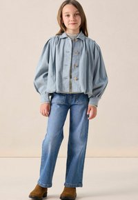 Une jeune fille se tient debout, portant une veste boutonnée bleu clair avec des manches bouffantes, un haut rayé, un jean bleu et des bottes marron sur un fond uni.