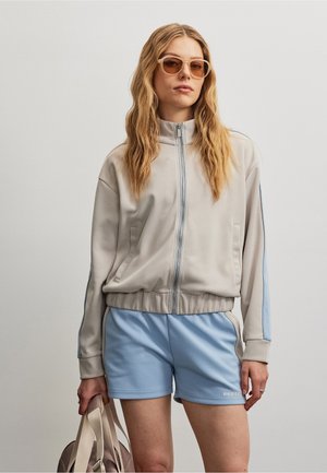 Frau mit langen, welligen Haaren, die eine beigefarbene Jacke mit Reißverschluss, hellblaue Shorts, eine Sonnenbrille trägt und eine beige Tasche vor einem einfarbigen Hintergrund hält.