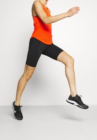 Ljus orange atletisk linne med texturerad tyg, matchat med svarta tajta shorts och svarta löparskor med vita detaljer.