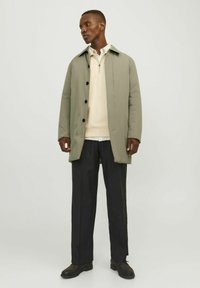 Abrigo impermeable verde claro con botones al frente, usado sobre un suéter crema y pantalones negros, con un corte relajado y cuello clásico.