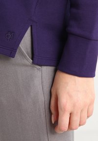 Top de algodón morado de manga larga con puños acanalados, que presenta aberturas laterales y un detalle de logo. Combinado con pantalones de mezclilla gris claro.