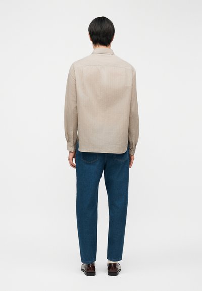 Άνδρας με κοντά μαύρα μαλλιά, φορώντας μπεζ μακρυμάνικο πουκάμισο, μπλε τζιν και καφέ παπούτσια, όρθιος με την πλάτη προς τα εμπρός σε απλό φόντο.