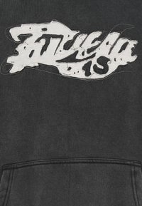 Must kapuutsiga pusa, millel on suur kulunud valge tikandembleem. Tekst sisaldab sõnu "Fitela 15", tekstureeritud puuvillase viimistlusega ja esiküljel taskuga.