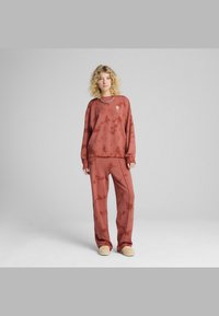 Rdeč majica s tie-dye vzorcem in usklajenimi hlačami iz mehke tkanine. Ima ohlapen kroj, okroglo ovratnik in subtilne oblikovalske podrobnosti na majici.