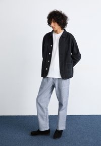Black cotton jacket with button closure and two front pockets, paired with light striped trousers and black shoes. Neutral background.