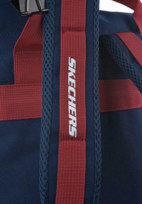 La mochila azul marino presenta correas rojas y acentos de malla. El logo "SKECHERS" se muestra de manera prominente en una etiqueta blanca. Se puede ver un tejido texturizado.
