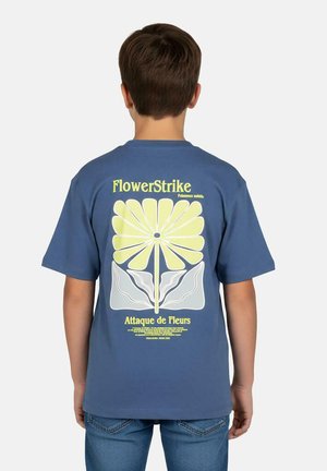 Kind draagt een blauw T-shirt met een grote gestileerde gele bloem en de tekst "FlowerStrike" en "Attaque de Fleurs" op de achterkant.