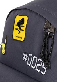 Borsa di tessuto grigia con una pattina gialla decorata da un logo nero, illustrazione di Pikachu e testo ricamato bianco "#0025". Chiusura con zip e tiranti neri.