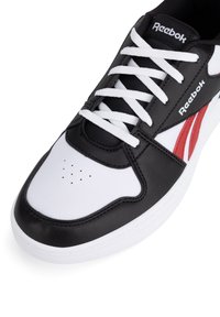 Fekete-fehér sportcipő sima bőr felsőrésszel, fehér fűzőkkel, perforált orral, valamint piros logóval és kiegészítőkkel.