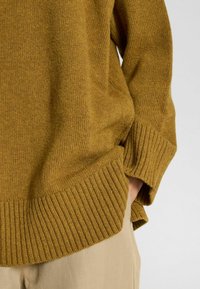 Sennepsfarvet strikket sweater med ribbede manchetter og kant, løs pasform og sideslidser forneden. Bæres over beige bukser.
