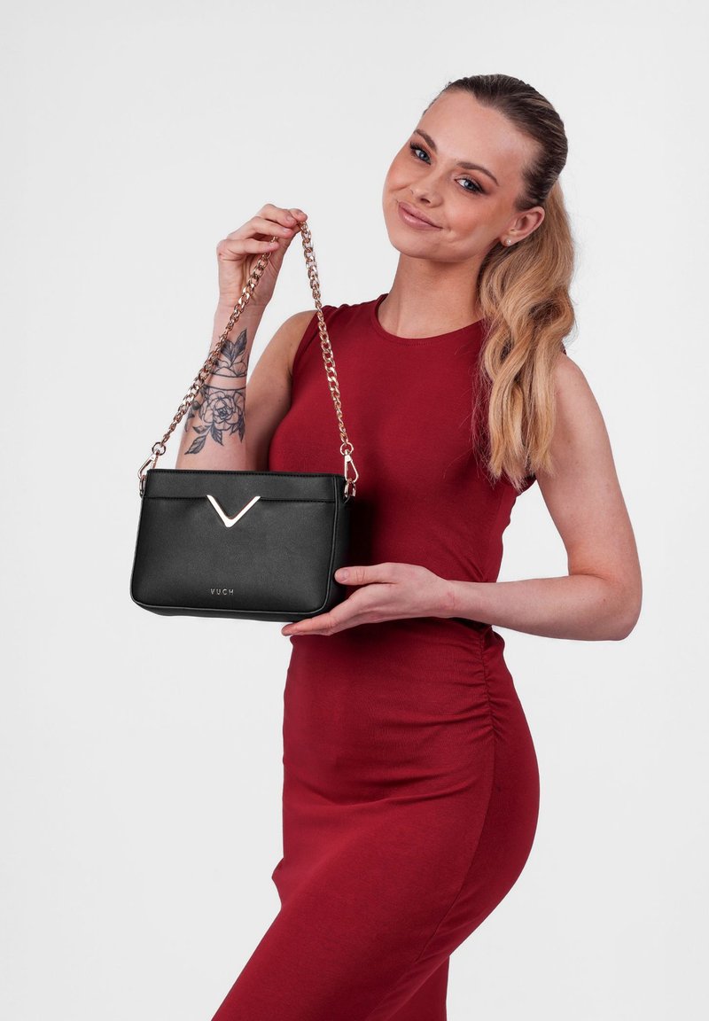 Černá umělá kožená kabelka s řetízkem; na přední straně má V-tvarovaný akcent a vyražené logo. Kompaktní obdélníkový design.