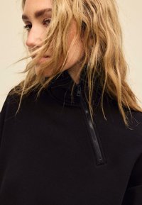 Črni pulover z visokim ovratnikom in zadrgo. Gladek tekstura tkanine z minimalističnim dizajnom, prikazuje posnetek obraza modela od blizu.