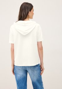 Hoodie blanc à manches courtes avec une coupe décontractée, présentant un design simple et un ourlet arrondi. Associé à un jean en denim bleu clair.