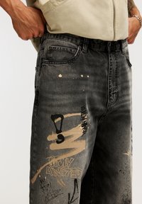 Svarta denimjeans med avslappnad passform, med abstrakta färgfläckar i beige och svart, synlig sömnad och framfickor.