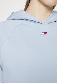 Jasnoniebieska bluza z kapturem, wykonana z miękkiego materiału, z kapturem na sznurku i małym haftowanym logo w czerwonych, białych i granatowych akcentach na klatce piersiowej.