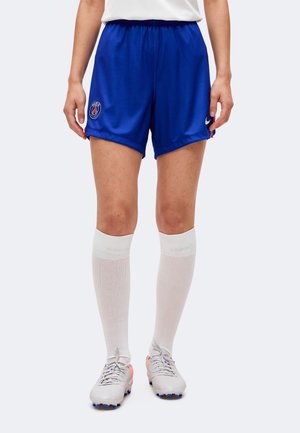 Personne portant un short de football bleu du Paris Saint-Germain, des chaussettes blanches montantes et des crampons blancs, debout devant un fond uni.