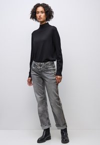 Pull à col roulé noir et jean denim gris avec accents latéraux. Le modèle porte des bottines noires. Coupe décontractée avec revers sur les jeans.