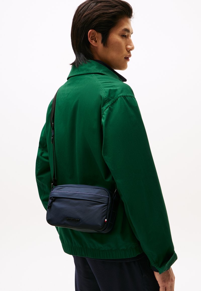 Námornícky modrá crossbody taška s uzáverom na zips a jemnými akcentmi. Hladká textura a kompaktný dizajn nosený proti zelenej bunde.