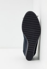 Semelle d'une chaussure plate noire avec des rainures horizontales et la taille 37 indiquée, positionnée verticalement contre un fond de coin blanc.