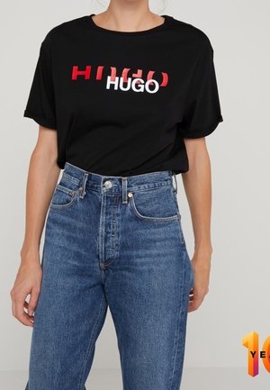 Musta puuvillainen T-paita, jossa punavalkoinen "HUGO"-logografiikka; rento istuvuus, pyöreä pääntie, yhdistetty korkeavyötäröisiin sinisiin farkkuihin.