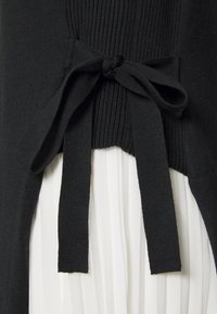 Tissu noir tricoté avec texture côtelée et détail de taille nouée, superposé à un tissu blanc plissé, mettant en valeur des styles contrastés.