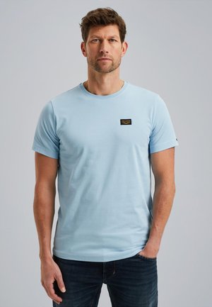 Light blue cotton t-shirt with short sleeves, round neckline, and a small black label on the chest. Paired with dark jeans.