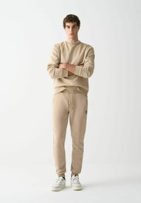 Sudadera beige y joggers a juego con puños elásticos, cintura con cordón y parche con logo. El modelo lleva zapatillas blancas.