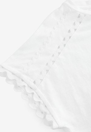 Biała koszulka z falbaniastymi brzegami i ozdobnymi wycięciami na rękawach, wykonana z miękkiego, lekkiego materiału.