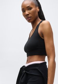 Femme athlétique avec une queue de cheval portant un brassière de sport noire et une veste nouée, debout devant un fond gris clair uni.