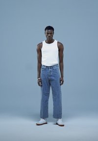 Valge tanktop, helesinised teksad ja valged jalanõud. Modell seisab ühevärvilise sinise tausta ees, rõhutades lõdvestunud lõiget ja peeneid tekstuure.