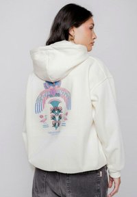 Sudadera crema con un gran gráfico en tonos pastel en la parte de atrás, que presenta patrones geométricos y diseños florales. Tejido suave, corte casual.
