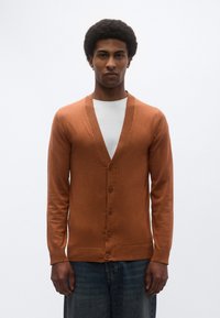 Καφέ μάλλινη ζακέτα με βαθύ V-λαιμόκοψη, ριμπ άκρα και πέντε κουμπιά, φορεμένη πάνω από λευκό t-shirt και συνδυασμένη με μπλε τζιν παντελόνι.