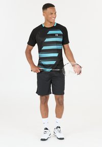 Svart träningsskjorta med blå diagonala ränder, tillsammans med svarta shorts. Modellen håller ett badmintonracket och har på sig vita strumpor och skor.