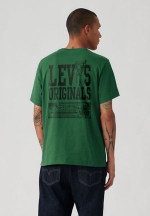 Man met tatoeages die een groen Levi's Originals T-shirt en een donkere spijkerbroek draagt, van achteren gezien waarbij het logo en de afbeelding op de achterkant van het shirt zichtbaar zijn.