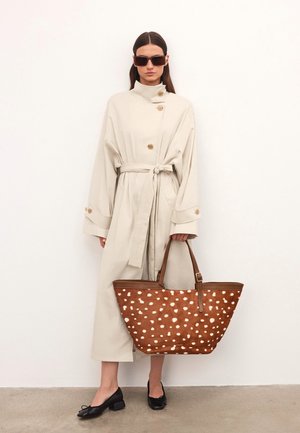 Trench beige con un cuello alto, detalles de botones y un cinturón atado, combinado con un bolso de mano marrón estampado que presenta diseños de puntos blancos. Bailarinas negras.