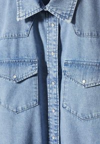 Camicia di denim azzurro chiaro con colletto all'americana, due taschini sul petto e bottoni a pressione argentati. Presenta una texture visibile e dettagli di cucitura.