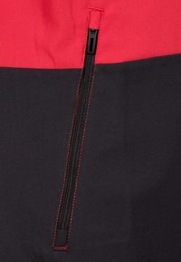 Mit Reißverschluss versehene Bekleidung mit einem rot-schwarzen Farbblockdesign. Strukturierten Stoff mit roten Nähakzenten entlang des Reißverschlusses für zusätzliche Details.