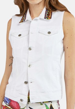 Donna che indossa un gilet bianco senza maniche abbottonato con colletto ricamato colorato e pantaloni con motivo floreale, mostrando un tatuaggio parziale sul braccio.