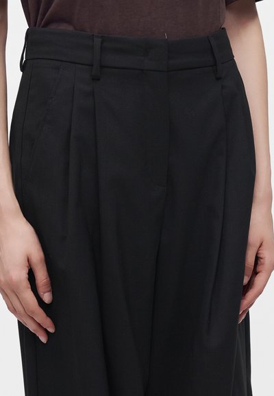 Personne portant un pantalon noir taille haute, ample, avec des passants de ceinture et des poches latérales, associé à un haut marron foncé.