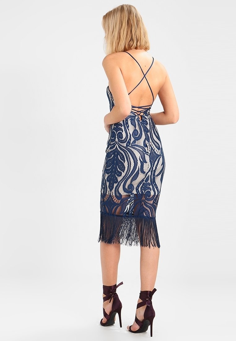 Robe en dentelle marine avec des motifs complexes, ourlet à franges et dos nu. Porte avec des chaussures à talons hauts ornées de rubans noués autour des chevilles.