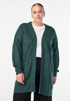 Groene, geribbelde gebreide cardigan met een open voorkant, lange mouwen en twee voorzakken, gedragen over een wit shirt en zwarte broek.