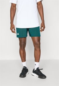 Shorts de sport vert foncé avec un motif texturé, arborant un logo blanc sur la jambe gauche. Associés à un t-shirt blanc et des baskets noires.